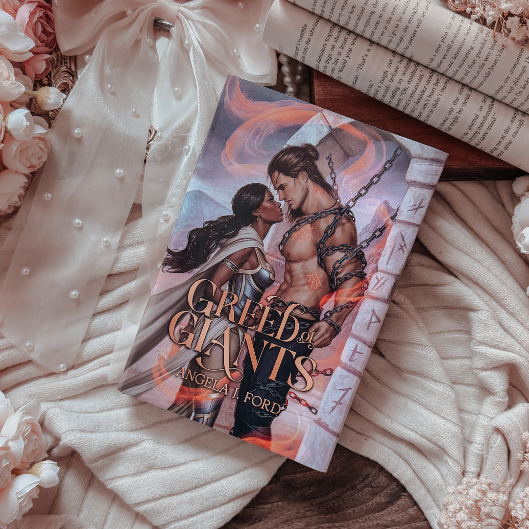 A book titled 'Greed: The Giants' by Angeliad Ford lies on a bed of soft cream-colored knit blankets, white pearls, pink roses, and an open book. The book cover features a romantic illustration of a muscular man and a woman with dark hair, bound by chains, with fiery orange and red tones.