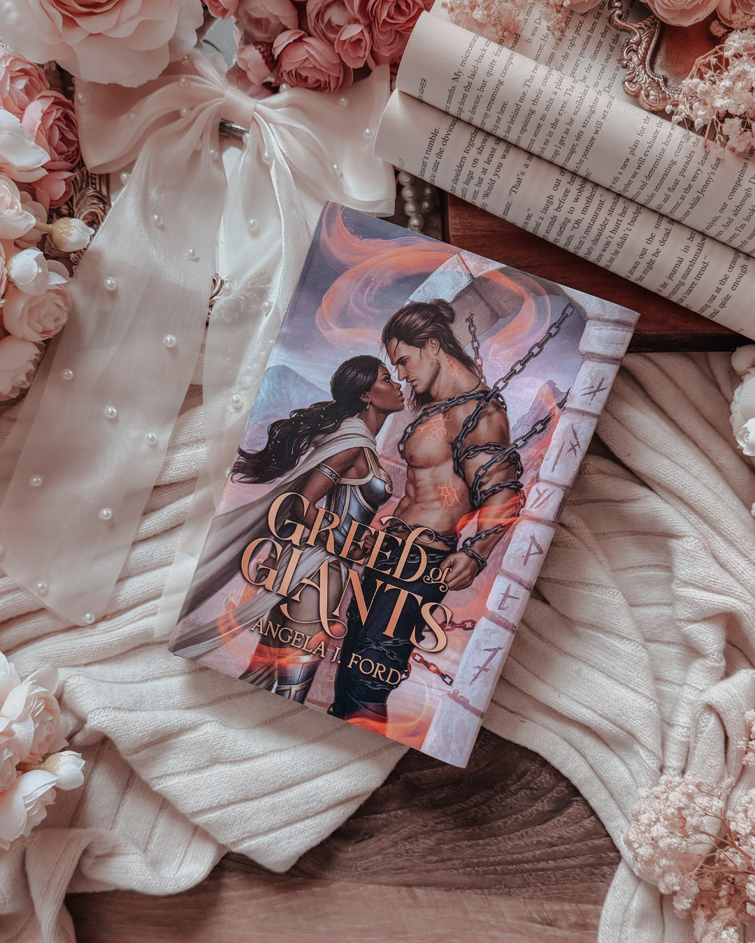 A book titled 'Greed: The Giants' by Angeliad Ford lies on a bed of soft cream-colored knit blankets, white pearls, pink roses, and an open book. The book cover features a romantic illustration of a muscular man and a woman with dark hair, bound by chains, with fiery orange and red tones.