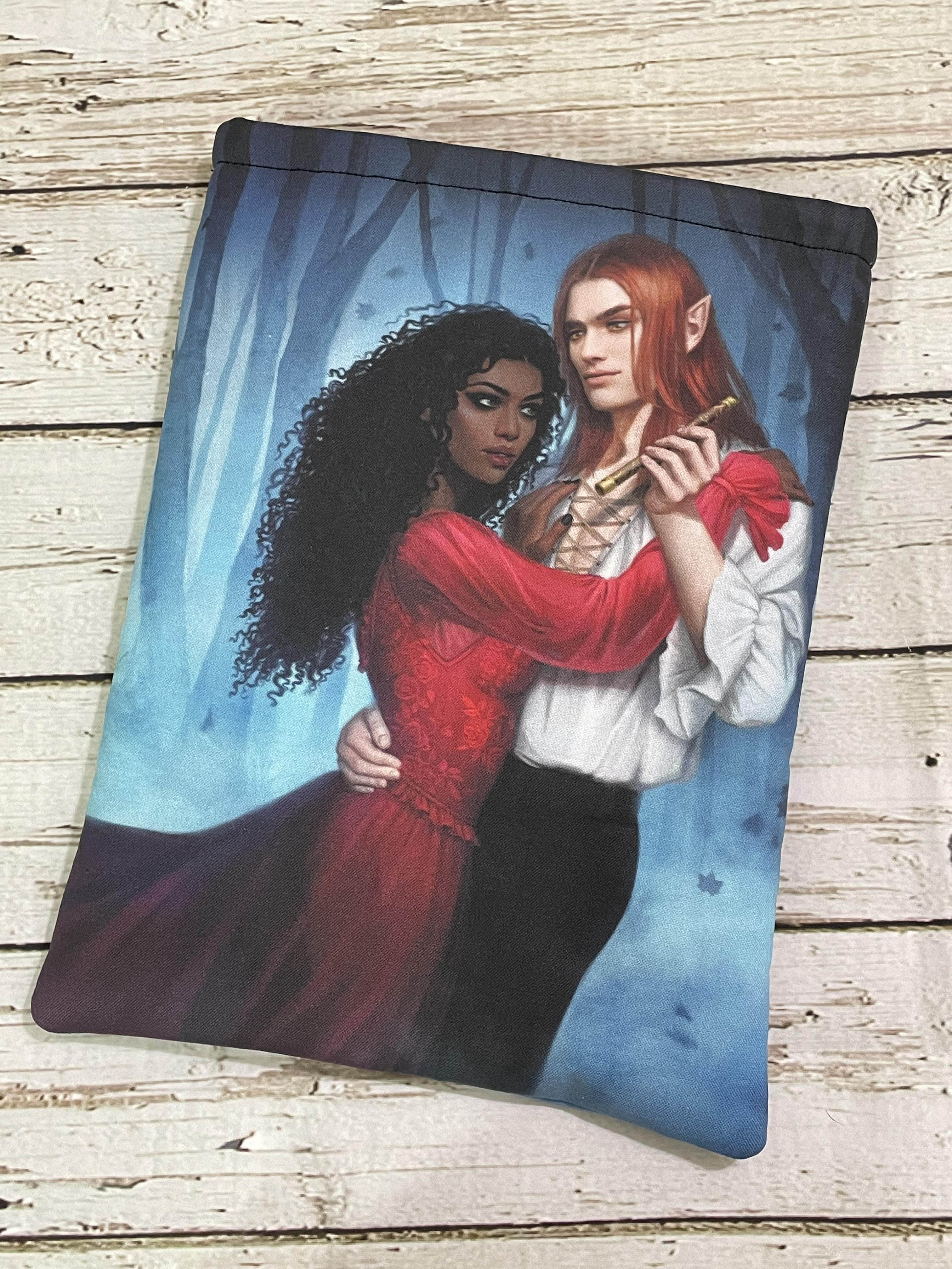 A printed pouch featuring an illustration of a dark-skinned woman in a red dress embracing a pale elf with long red hair, dressed in white, standing together in a blue, misty forest.